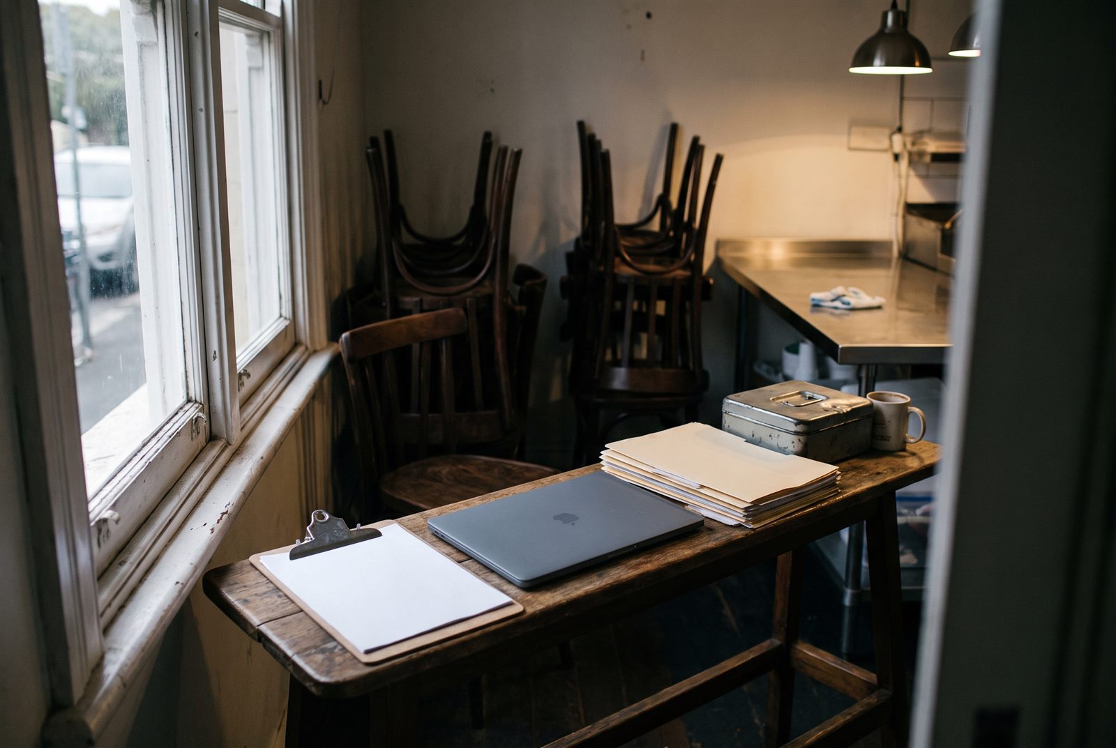 Back of house corner of an Australian cafe after closing, with a clipboard of rostering sheets and a closed laptop on a narrow bench