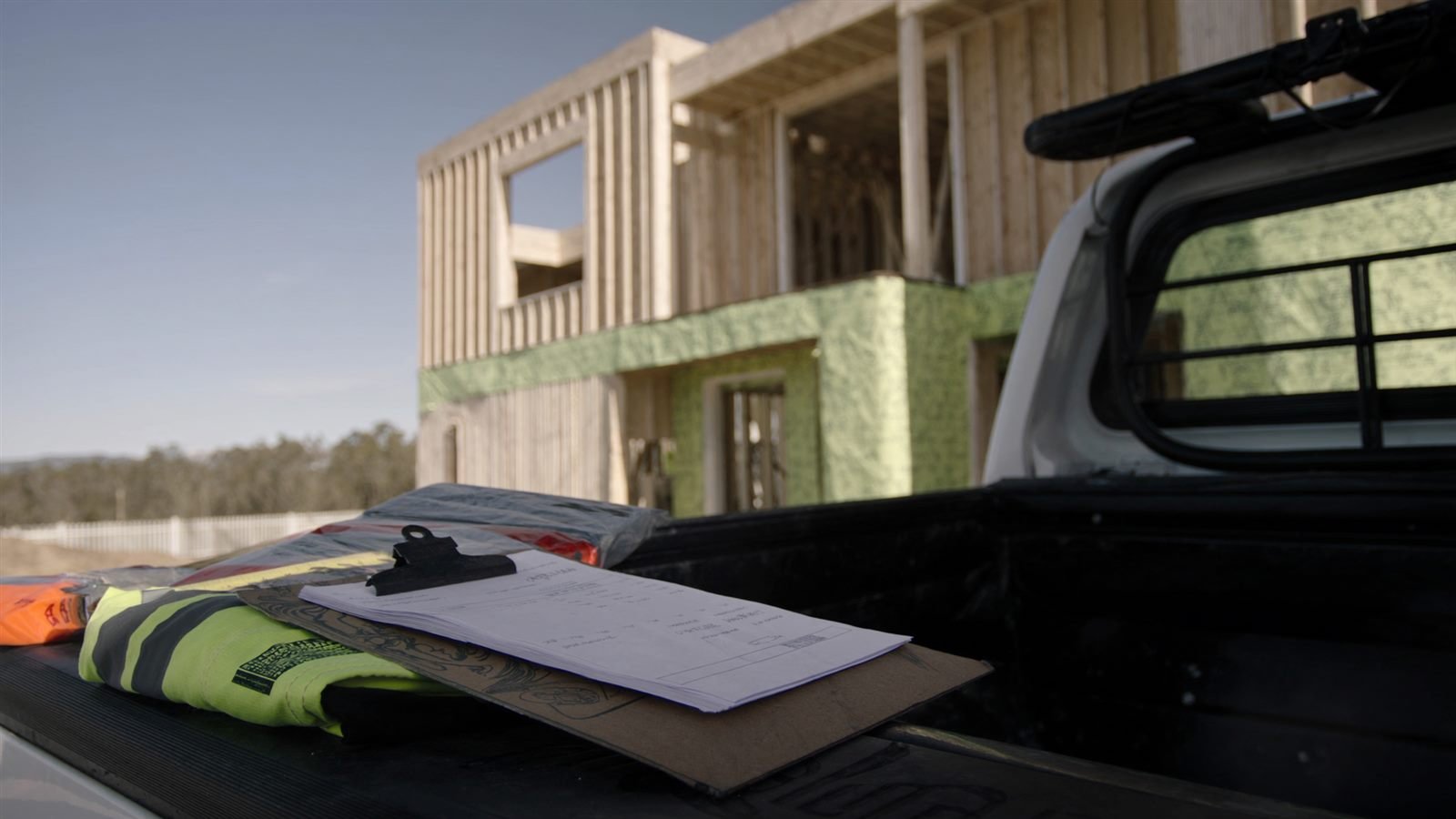 Clipboard of paper subcontractor invoices on the tray of a white ute parked at an Australian house frame building site, beside a hi-vis vest and tape measure