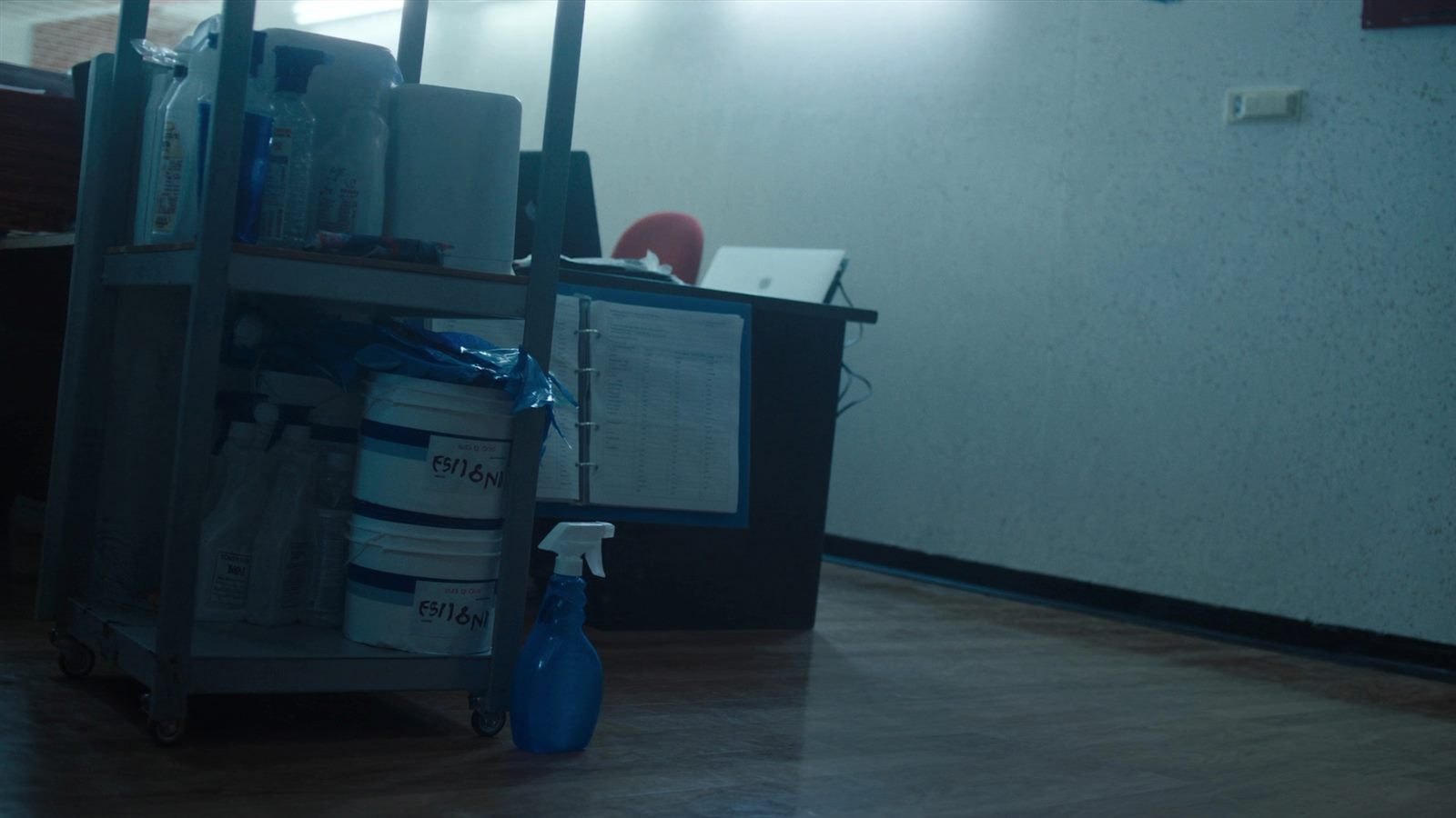 Cleaning supplies trolley with buckets and spray bottles in a small cleaning business office, beside a desk with a ring binder of contractor timesheets and a laptop