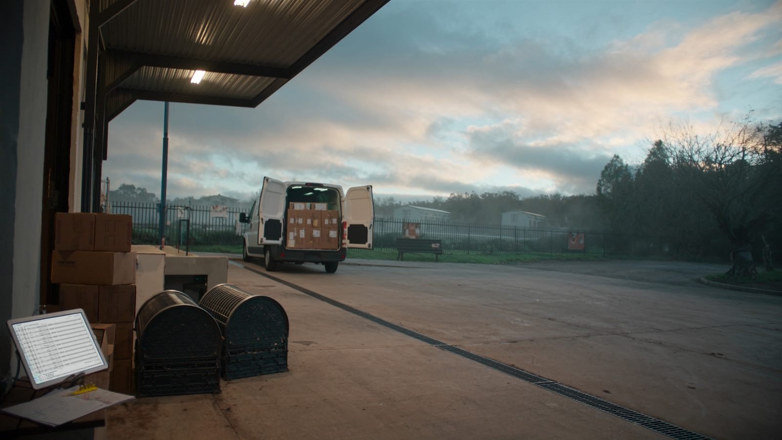 Loading dock of a small Australian courier depot at dawn, with stacked cartons, a tablet showing a spreadsheet and a white van loaded with boxes, rear doors open