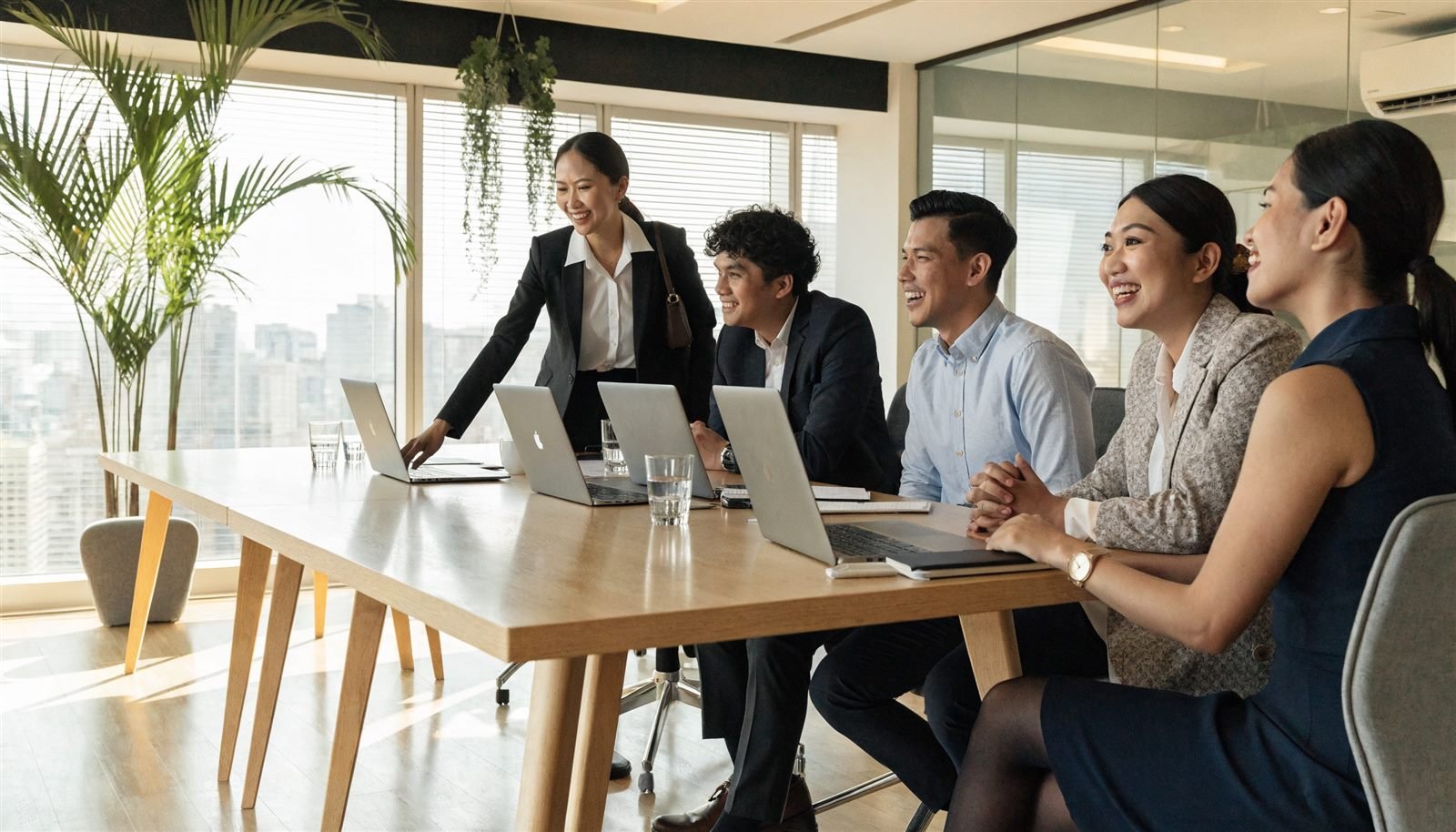 Five accountants in business attire laughing around a long timber boardroom table with laptops in a bright Manila office with palms and a city view