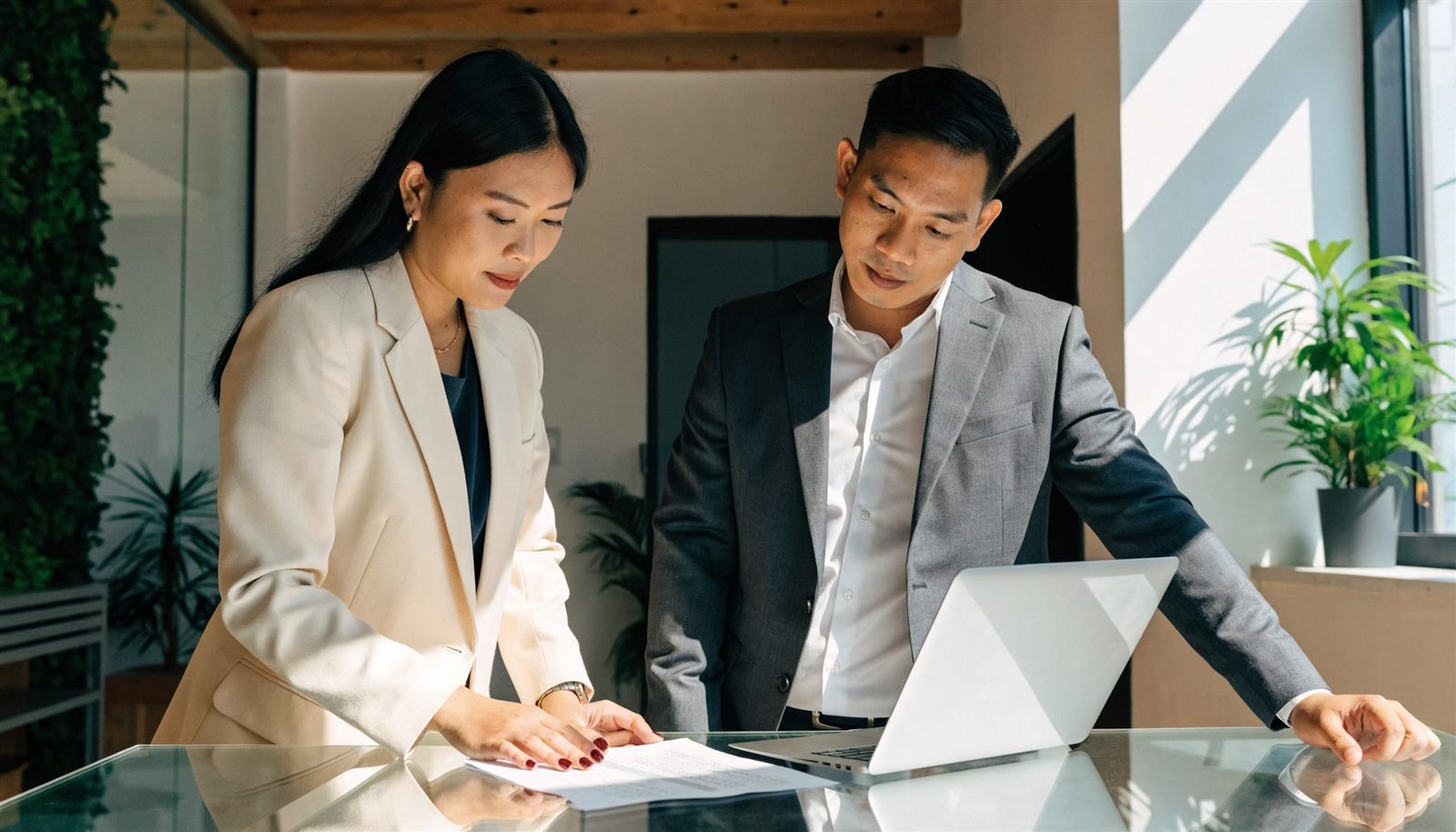 Two accountants in business attire standing at a glass meeting table in a sunlit Manila office, reviewing a printed pay run beside an open laptop